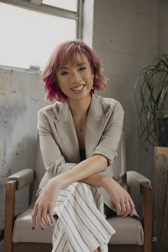 Image of Asian Australian girl with pink hair in a beige blazer and white pants, sitting on a chair, leaning forward, smiling at camera.
