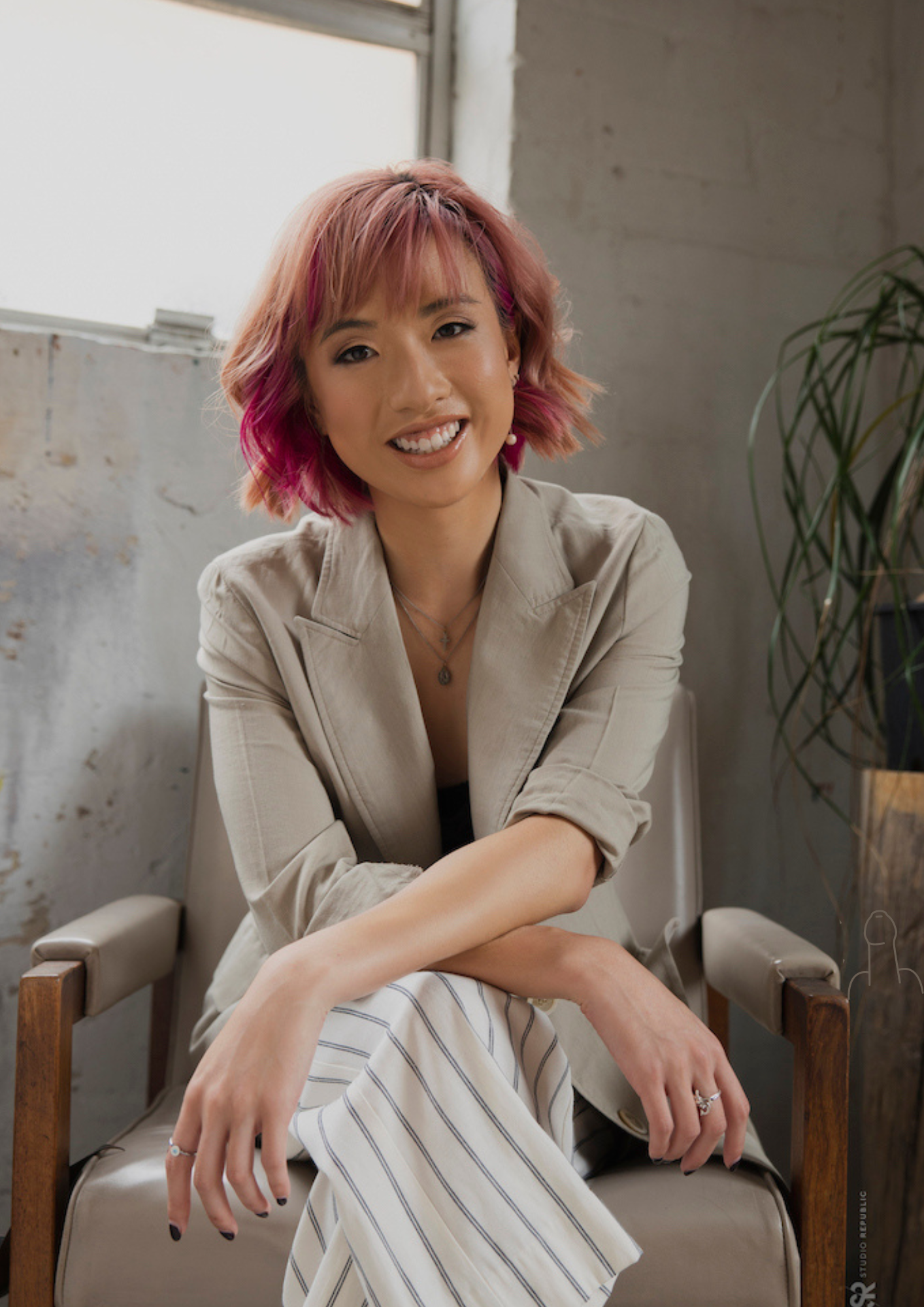 Image of a Chinese Australian woman with pink hair in a grey blazer, sitting relaxed and smiling on a chair.